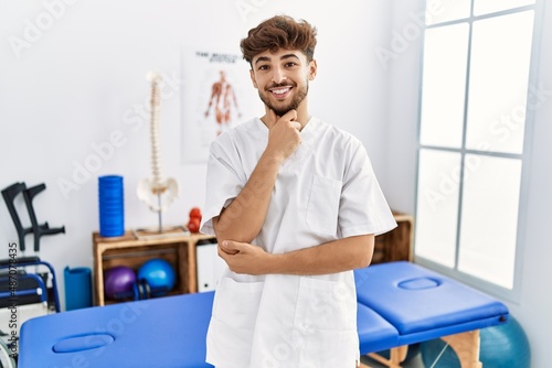 Young arab man working at pain recovery clinic looking confident at the camera smiling with crossed arms and hand raised on chin. thinking positive.