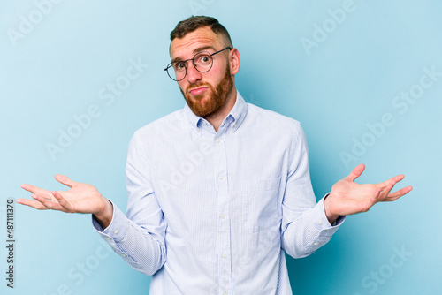 Young caucasian man isolated on blue background doubting and shrugging shoulders in questioning gesture.