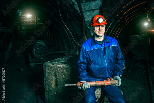 A tired miner in an old coal mine sits on a broken trolley. A miner in a blue protective suit with an orange helmet on his head holds a jackhammer. Copy space.