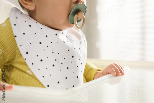 Cute little baby wearing bib in highchair indoors, closeup