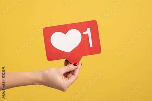 Support interesting web content, push heart button. Closeup of woman hand holding social media like icon, emoji sign to follow subscribe blog. Indoor studio shot isolated on yellow background.