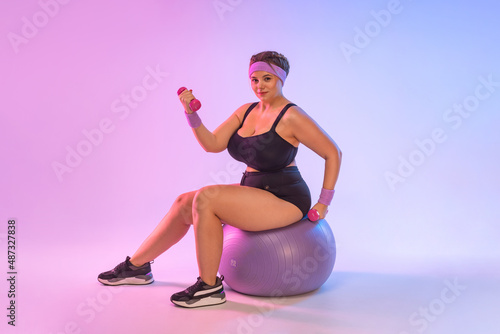 Size plus fitness model, athletic trainer instructor woman in home gym sit on fitball isolated on pink background. Workout sport concept