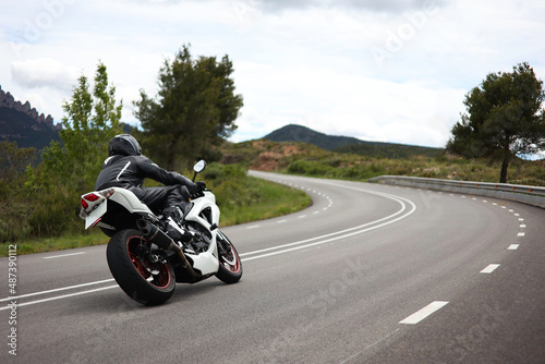 Biker in a black uniform driving a white sports motorcycle on a curvy road in the middle of a green meadow with trees and with mountains in the background on a cloudy day