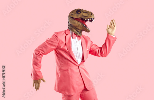 Funny man in rubber dinosaur mask dancing and having fun in the studio. Happy lizard headed guy in stylish funky vibrant pink party suit doing Egyptian dance moves isolated on pink colour background