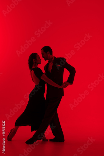 Dark silhouettes of tango dancers couple on red background. Man in classic suit and woman in elegant dress in dancing movement.