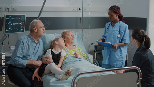 Nurse doing consultation with sick woman while family visits her in hospital ward bed. Medical assistant talking to elder woman and people about illness. Mother, kid and man in visit