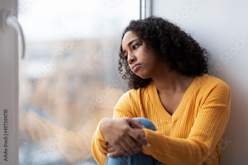 Depressed young African American woman sitting near window, looking outside on street, feeling hopeless at home