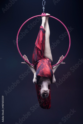 Circus performer woman in red dress doing tricks on red Lyra isolated on black background.
