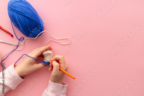 Children's hands in the process of crocheting toys from blue and beige yarn. Pink background. Needlework, hobbies, craft training. The development of fine motor skills. Flat lay, copy space.