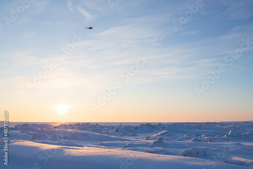 The helicopter is flying in the sky over the frozen sea in the Far North in the Arctic. Travel and tourism in the polar region. Cold winter weather. Ice hummocks and snow. The sun is above the horizon