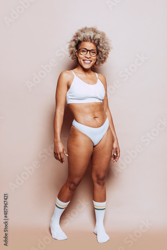 Cheerful beautiful afro latin american woman in white underwear and eyeglasses isolated on beige background at studio. Stretch marks and cellulite on the skin. Real people, real body, natural beauty.