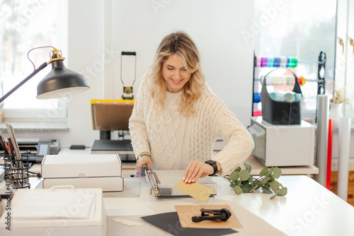 Professional smiling blond woman in sweater worker using printer cutter in workshop. Print office. Carving sheets paper 