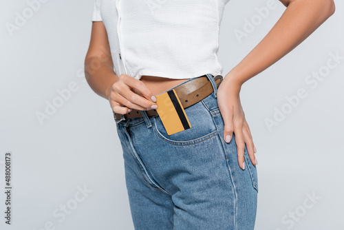 cropped view of young woman putting credit card with cashback in pocket on jeans isolated on grey.
