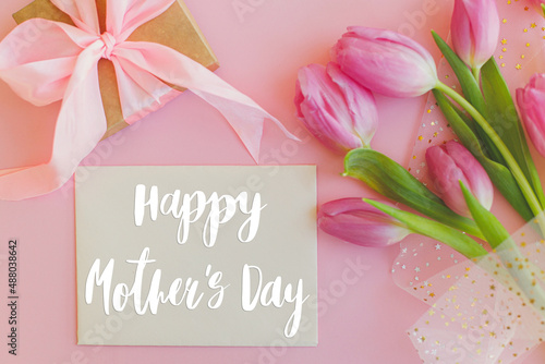 Happy mothers day text on greeting card, pink tulips bouquet and gift box on pink background. Stylish greeting card. Happy Mother's Day, gratitude and love to mom. Handwritten lettering