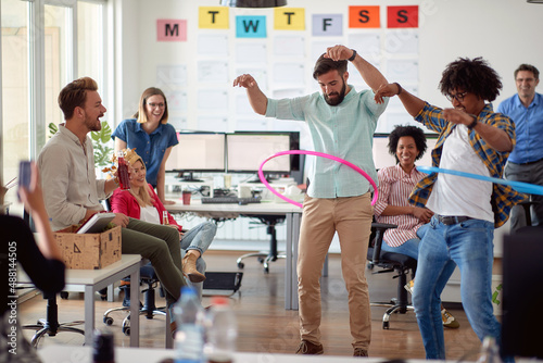 Two male employees is playing with hula-hoop in the office with their colleagues. Employees, job, office