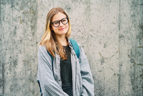 Outdoor portrait of young teenage kid girl wearing glasses and backpack, posing on grey wall background