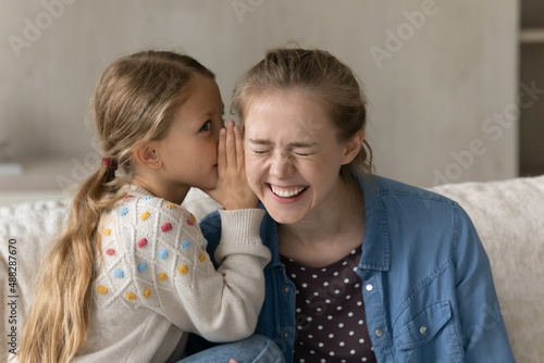 Cute kid telling secret to happy laughing elder sister, nanny, daycare teacher, whispering in ear. Pretty loving daughter girl and young mom enjoying friendship, having fun, talking at home