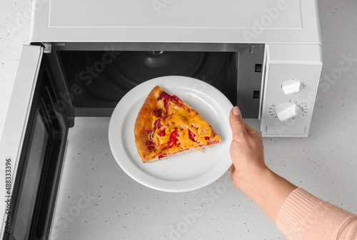 Woman putting plate with piece of pizza into microwave oven on table, closeup