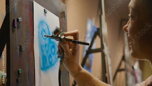 Woman artist learns to paint with airbrush with acrylic dye, paper and easel. Indoors. Concept modern art, airbrushing, aerography, draw spraying picture create sprayer drawing graffiti aerosol spray