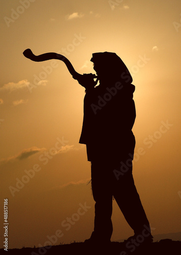 Silhouette of the entire body of a Jewish man wearing a tallit and tefillin and blowing a long shofar made from the horn of a kudu antelope at sunrise during morning prayers in Israel.