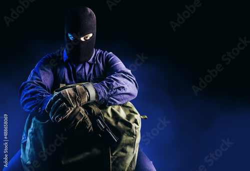 Photo of robber in mask, overalls, gloves, gun sitting and holding bag on dark blue background.