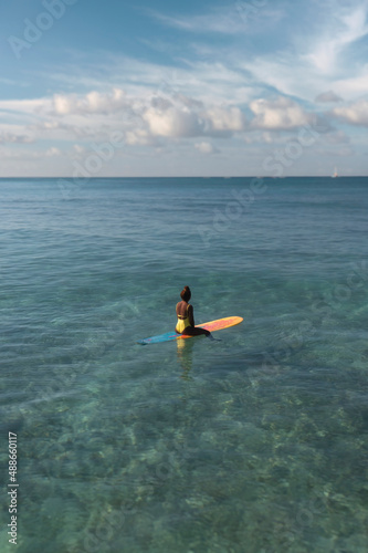 Charming curly African-American dark-skinned young woman, professional surfer sits on a long surfboard in the ocean, aerial shot from above