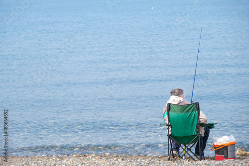 Man fishing on beach, one single male sitting on chair with fishing rod on coast with calm blue sea background. Beautiful minimalist concept with area for ad or advertisement text.