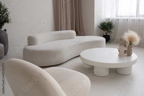 Large living-room with white comfortable sofa and armchair and round table with group of handmade vases with dry wildflowers