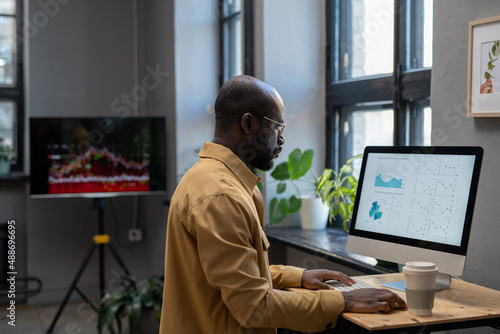 Young modern economist in casual clothes working with financial data in front of computer while sitting by desk in office