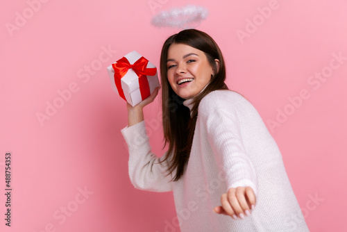 Satisfied angelic woman with nimb over her head, throwing present box, congratulating with holiday, wearing white casual style sweater. Indoor studio shot isolated on pink background.