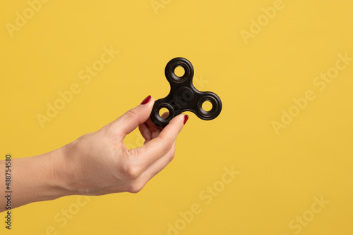 Closeup side view profile portrait of woman hand holding black fidget spinner, stress relieving toy. Indoor studio shot isolated on yellow background.