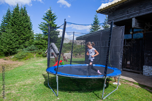 Happy little girl jump on trampoline in backyard. Child fun outdoor. Summer day. Blue sky. Nature landscape in background.