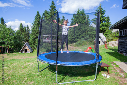 Happy little girl jump on trampoline in backyard. Child fun outdoor. Summer day. Blue sky. Nature landscape in background.