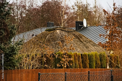 Blaszany dach domu z kominem i antenami , na pierwszym planie domek ( altana , wiata) ogrodowy pokryty strzechą , za płotem ze sztachet i ogrodzeniem z siatki stalowej .