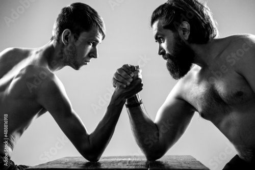 Heavily muscled bearded man arm wrestling a puny weak man. Arms wrestling thin hand, big strong arm in studio. Two man's hands clasped arm wrestling, strong and weak, unequal match. Arm wrestling