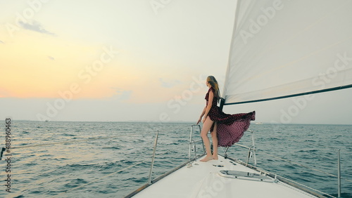 Side view of attractive woman in dark dress, travelling on yacht at sunset. Young slim girl standing on bow of yacht, raising up hand, watching seascape while sun setting down. Concept of lifestyle