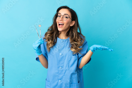 Woman dentist holding tools over isolated on blue background with shocked facial expression