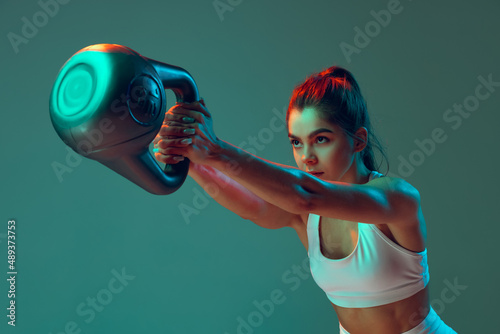Portrait of sportive woman workout, doing exercises with sports equipment isolated on green studio background in neon light. Sport, action, fitness, youth concept.