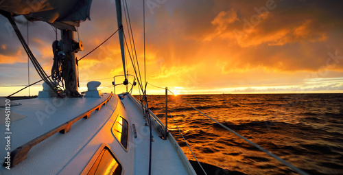 White yacht sailing in an open sea at sunset. A view from the deck to the bow, mast, sails. Epic cloudscape. Dramatic sky with glowing golden clouds after the storm. Racing, sport, leisure activity