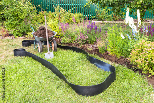 Two-wheeled wheelbarrow loaded with fertile soil, shovel and bender board before edging of the flower bed in the summer garden