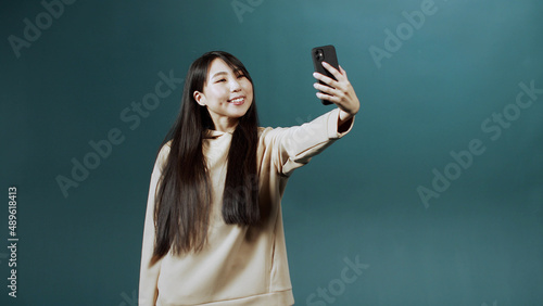Pretty girl in beige hoodie looks into the screen of new mobile phone, makes selfie, shows her tongue