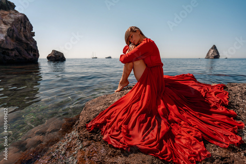 Beautiful sensual woman in a flying red dress and long hair, sitting on a rock above the beautiful sea in a large bay.