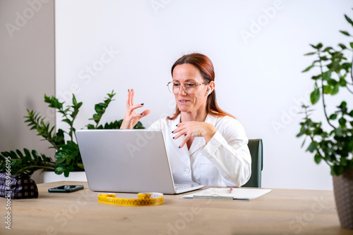 Portrait of female nutritionist in medical gown using laptop for online consultation via video chat with her patient while sitting in her office. Dietitian with laptop giving online weight loss