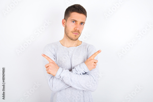 Serious young caucasian man wearing grey sweater over white background crosses hands and points at different sides hesitates between two items. Hard decision concept