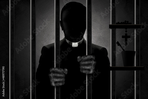 A Catholic priest identified by his clergyman collar is seen in silhouette behind bars with his filthy hands gripping the bars. He is in shadows and his face is in darkness.