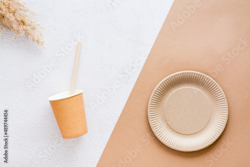 The concept of eco-friendly utensils for food. Cardboard cup, drinking straw, plate and ear of grass on plaster and beige background. Top view.