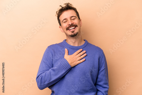 Young caucasian man isolated on beige background laughs out loudly keeping hand on chest.