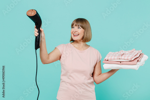 Elderly fun happy housewife woman 50s in pink t-shirt doing housework hold pile of clothes use steamer ironing isolated on plain pastel light blue background Housekeeping cleaning tidying up concept.