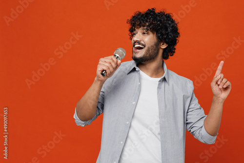 Young bearded Indian man 20s years old wears blue shirt sing song in microphone dance sing song fooling around have fun gesticulating hands enjoy isolated on plain orange background studio portrait.