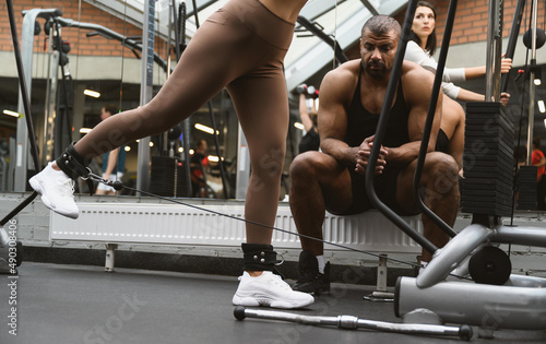 A young woman performs an exercise in a crossover machine. Personal trainer afro american man teaches how to do the exercise correctly